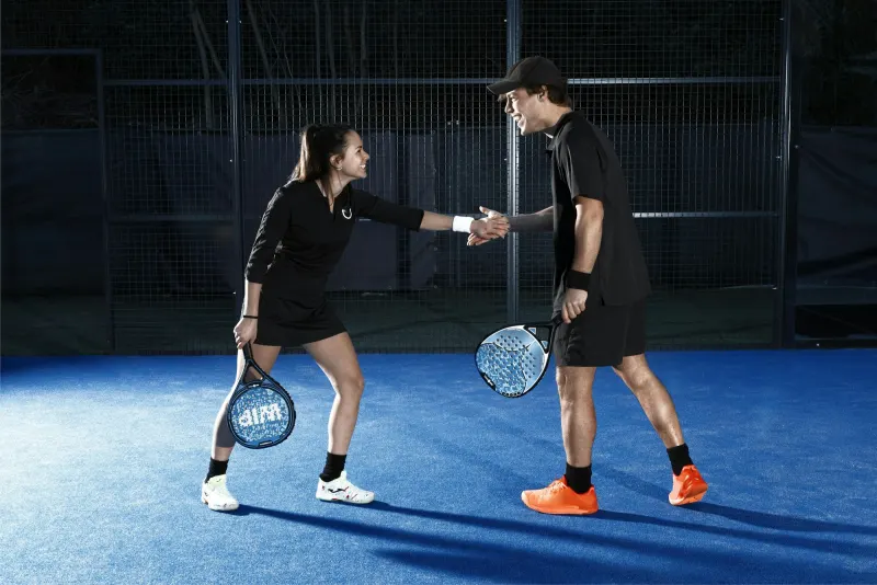 Two padel players shaking hands across a blue court at night